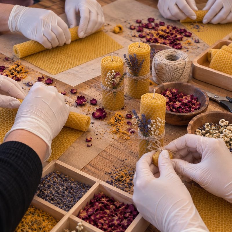 Gloved hands rolling beeswax candle sheets at a workshop table scattered with dried flowers, lavender and twine